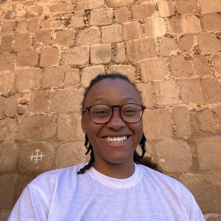 A woman with glasses and braids standing in front of a brick wall.